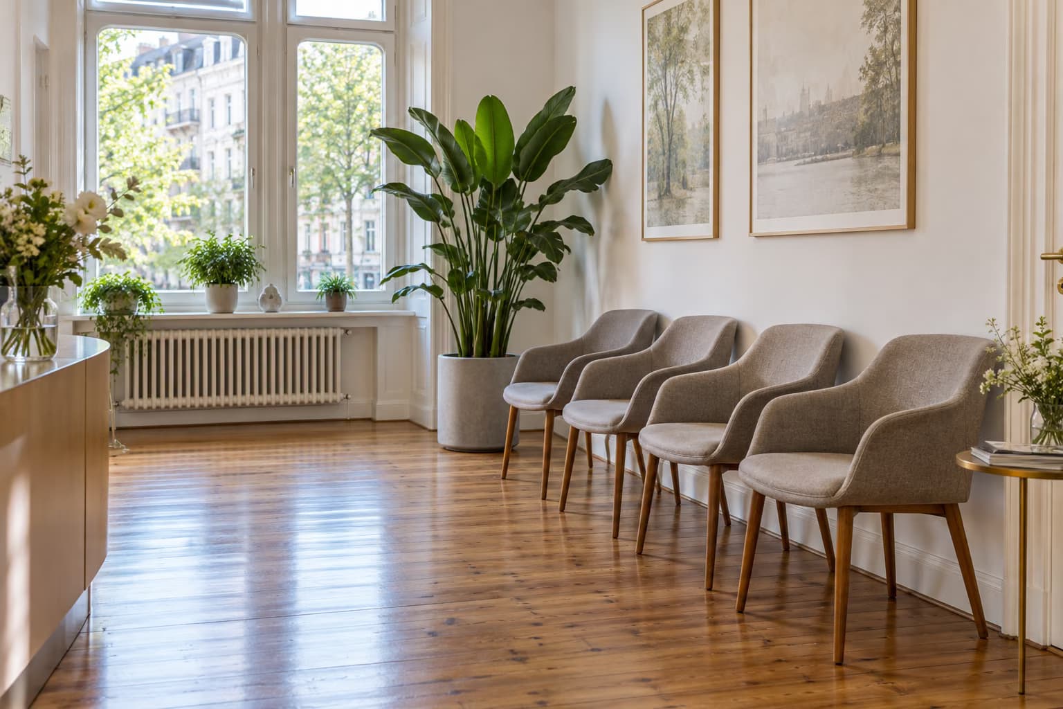 Uma sala de espera luminosa com uma fila de cadeiras estofadas cinzentas, uma grande planta em vaso e janelas altas viradas para uma rua arborizada.