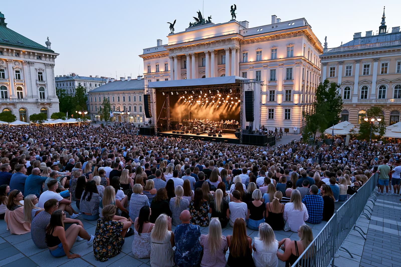 Ao entardecer, uma multidão se reúne em uma praça histórica de Berlim, onde um palco de orquestra ao ar livre é iluminado contra grandes fachadas.