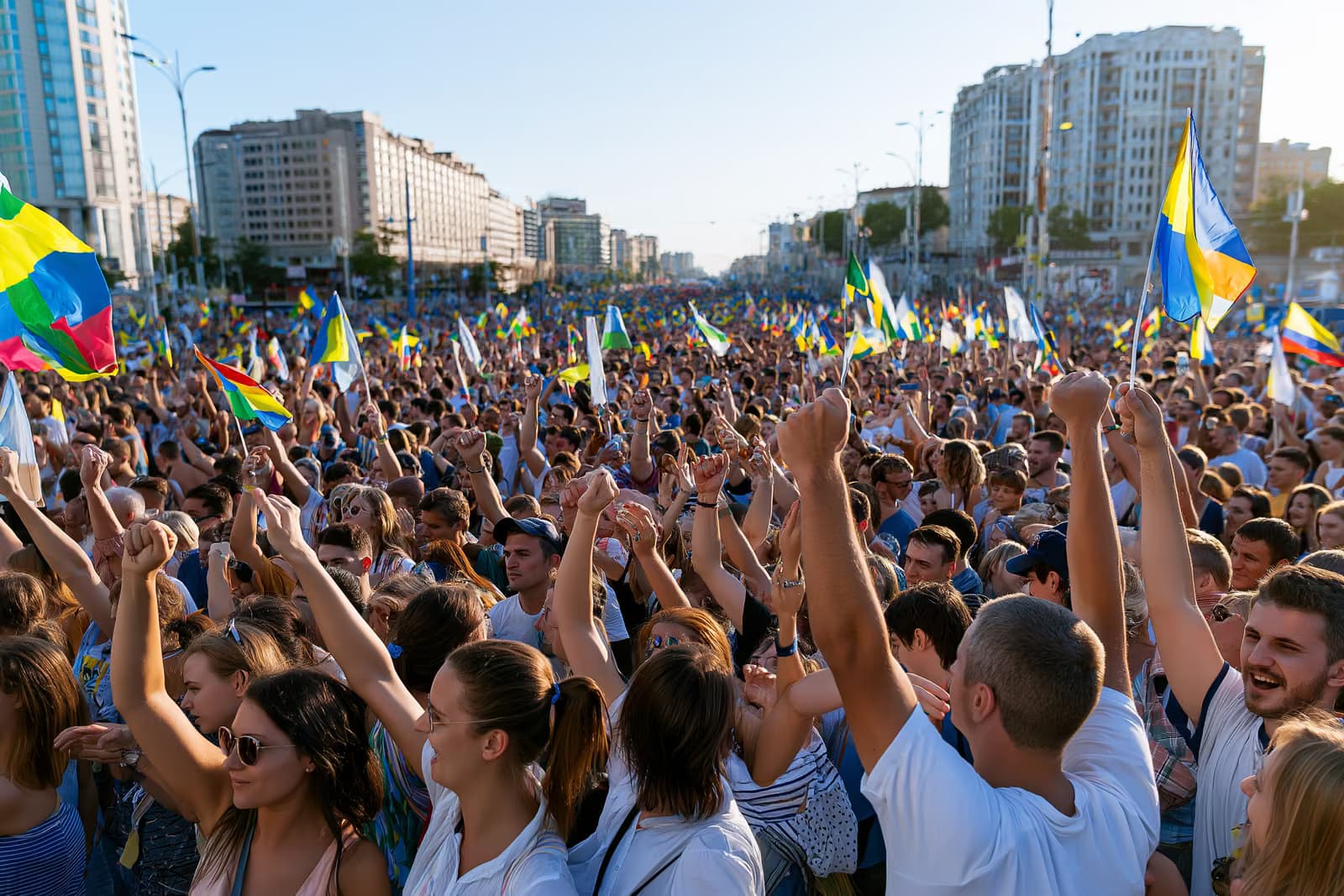 Uma enorme multidão lota uma rua da cidade sob um céu claro de verão, agitando bandeiras coloridas.