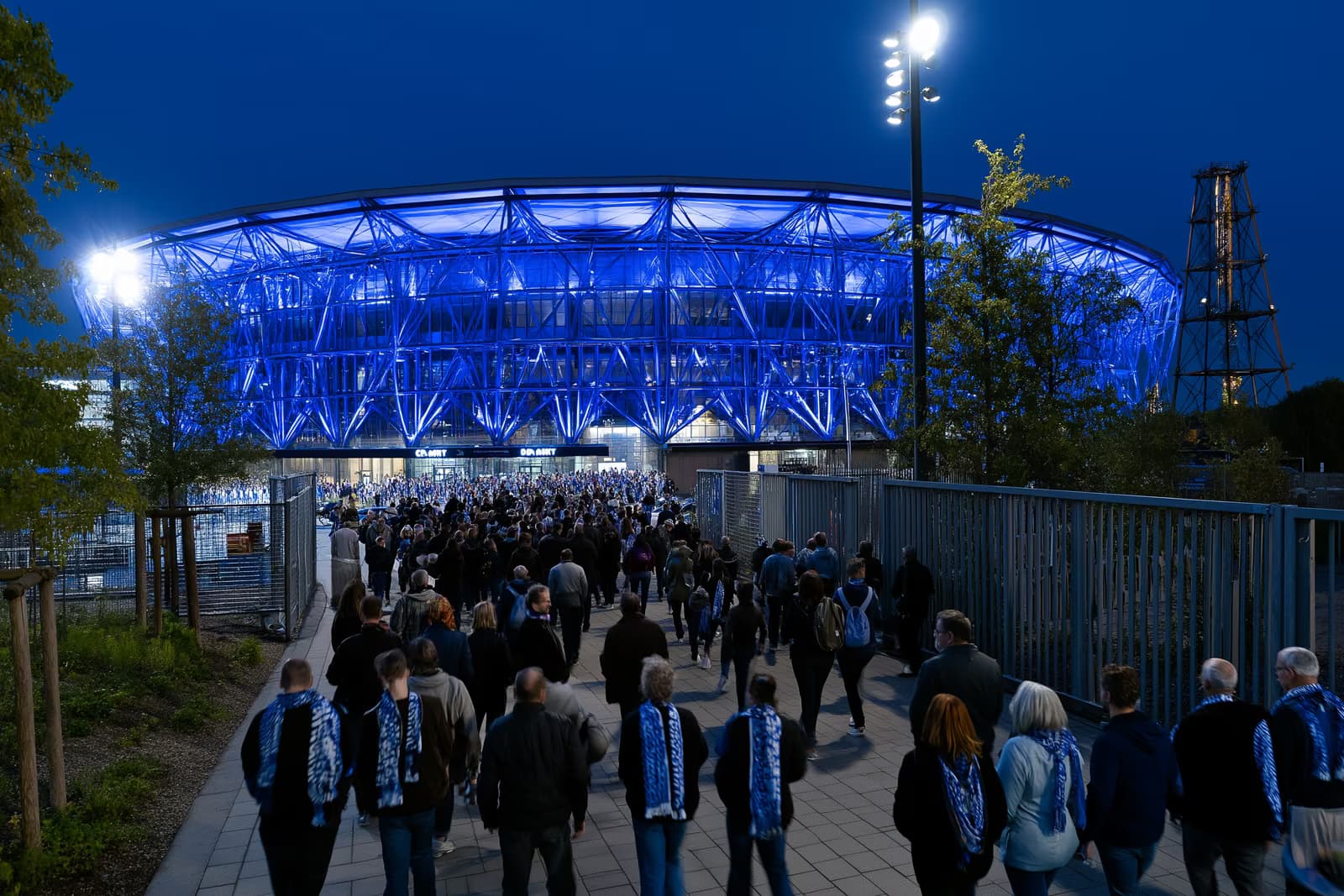 Um estádio de futebol na região do Ruhr brilha em azul real à noite, enquanto torcedores com cachecóis se dirigem aos portões após a mudança do Gosens para o Schalke.
