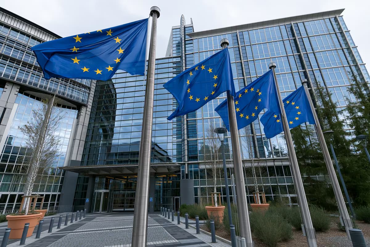 Bandeiras da União Europeia hasteadas em mastros em frente a um moderno edifício de vidro.