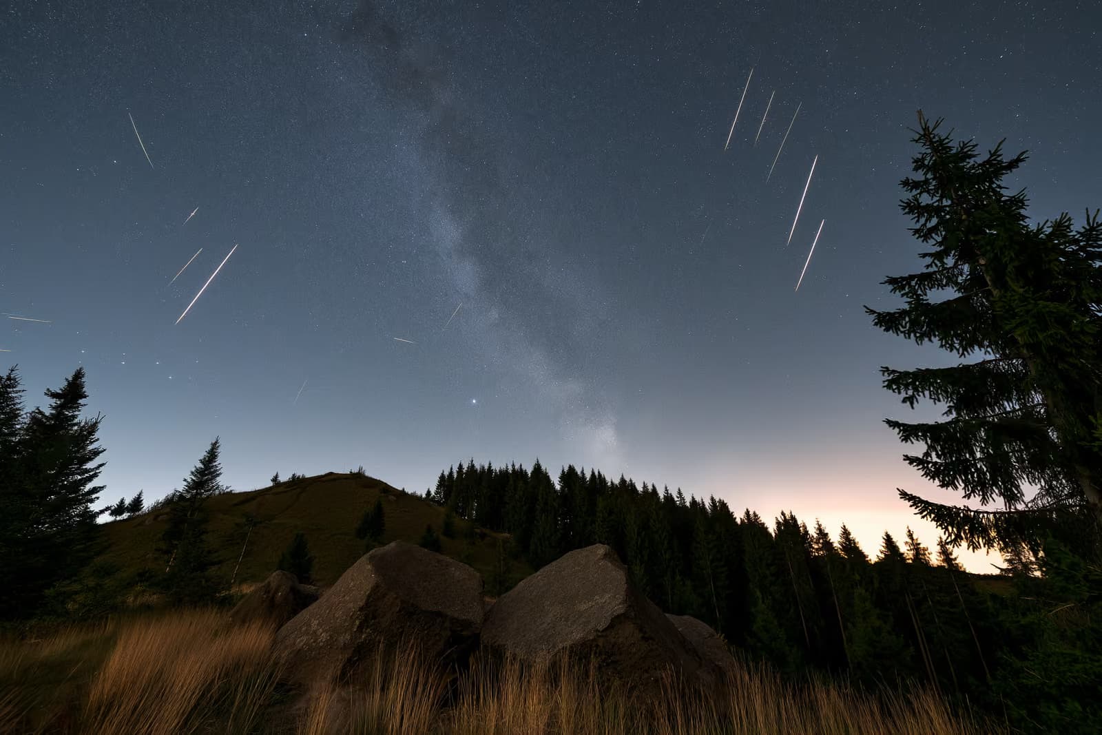 Um céu noturno sobre uma paisagem rural escura da Alemanha, com rastros de meteoros e uma tênue faixa de estrelas acima.